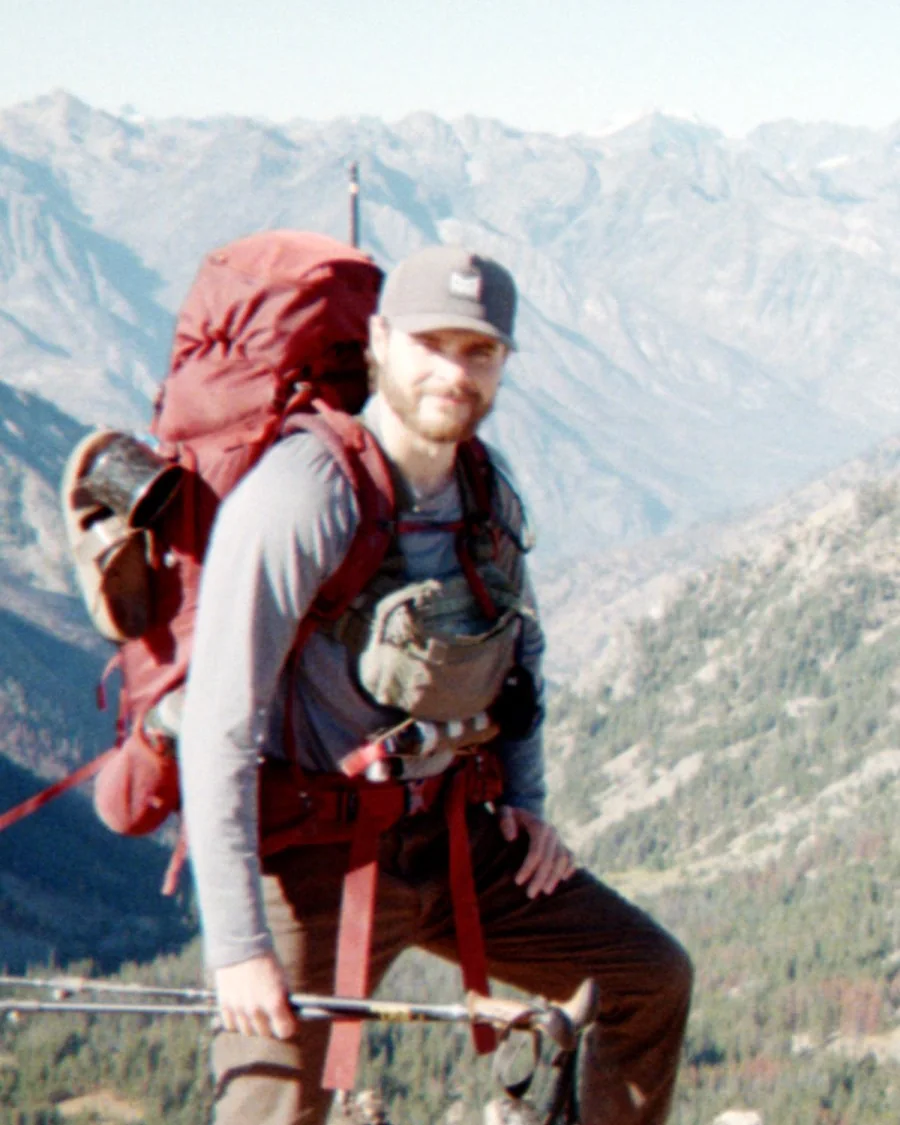Conner Crowe with a full pack and trekking poles on a ridge, smiling at the camera, the North Cascades behind him.