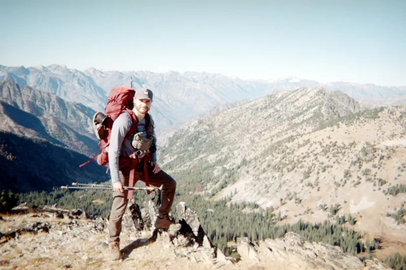 Conner Crowe with a full pack and trekking poles on a rocky ridge above a forested valley, with the North Cascades behind him.